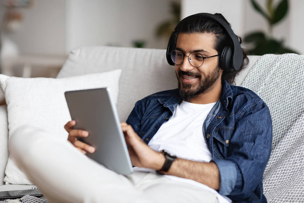 Guapo indio en auriculares inalámbricos relajante con tableta digital en casa - Foto, Imagen