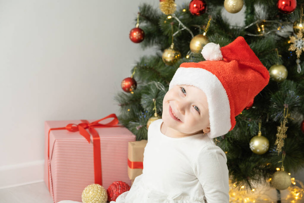 Nahaufnahme Porträt kleines Mädchen mit Weihnachtsmütze lacht in der Nähe des Weihnachtsbaums. - Foto, Bild