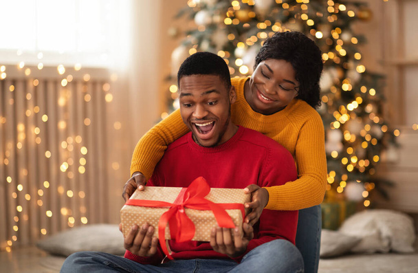Έκπληκτος αφροαμερικανός νεαρός σύζυγος κρατά κουτί με δώρο και κόκκινη κορδέλα - Φωτογραφία, εικόνα