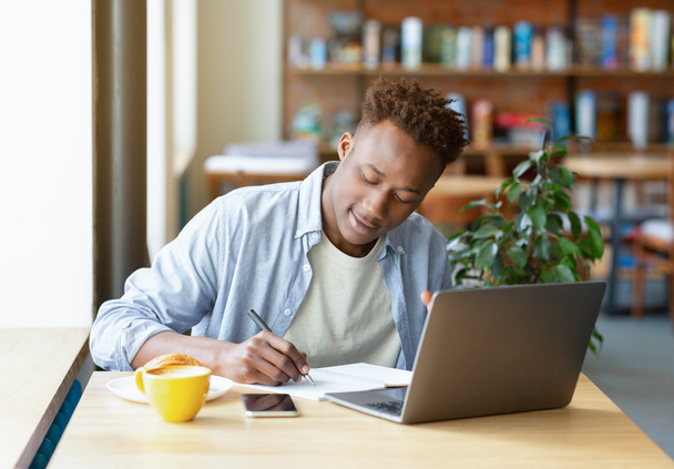 Pozytywny czarny człowiek za pomocą laptopa do pracy online lub badań, pisanie w notebooku, robi projekt w przytulnej kawiarni miejskiej - Zdjęcie, obraz