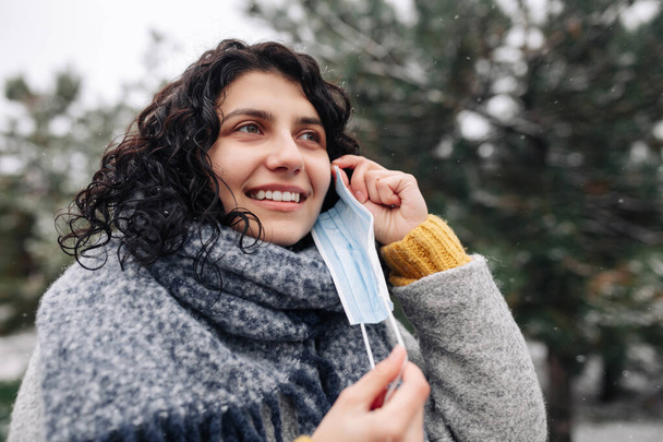 Молода жінка знімає медичну стерильну маску в зимовому засніженому парку в холодний морозний день. Коронавірус поширює запобігання спалаху пандемії дружби. Особисте захисне обладнання та концепція здоров'я
 - Фото, зображення