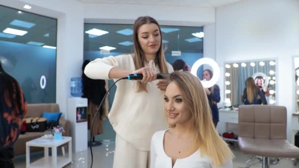 Jeune femme brune coiffeuse fait coiffage sur les cheveux longs de la fille blonde - Séquence, vidéo