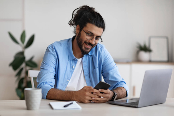 Western freelancer guy sitting at desk at home office and using smartphone - Photo, Image