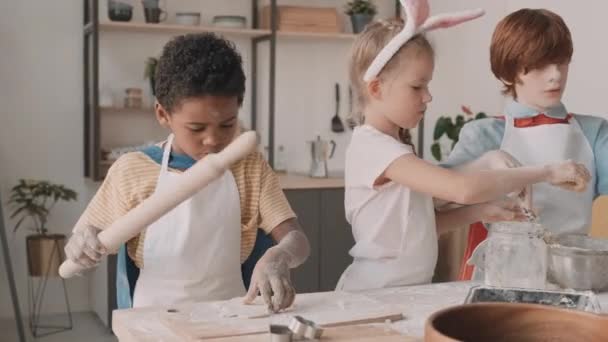 Waist up of African school boy rolling out raw pastry with wooden rolling pin on cutting board, girl with headband with bunny ears and red-haired Caucasian male friend making more dough - Footage, Video