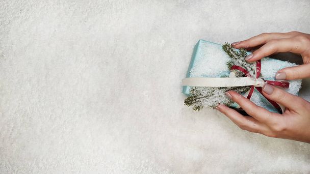 Female hands in snowflakes with a handmade Christmas gift box with red and white ribbon and snow. Top view with copy space - 写真・画像