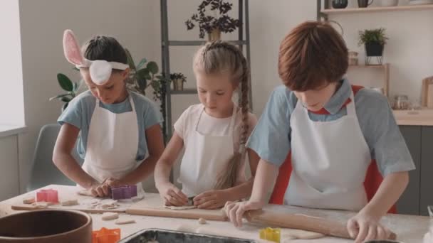 Waist up of red-haired boy rolling out dough with wooden rolling pin on table, Caucasian and Mixed-race school girls using cookie cutters. Children in aprons standing in kitchen, cooking - Footage, Video
