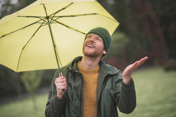 Young bearded man standing with yellow umbrella under the rain - Фото, зображення