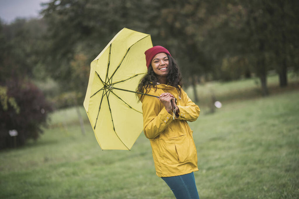 Cute mulatta with yellow umbrella looking cheerful - Zdjęcie, obraz