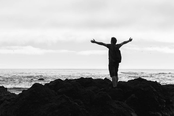 Denizde bir uçurumun tepesinde elleri havada duran bir adamın silueti. Özgürlük iyi hissettiriyor ve seyahat macera tatili konsepti, siyah beyaz fotoğraf - Fotoğraf, Görsel
