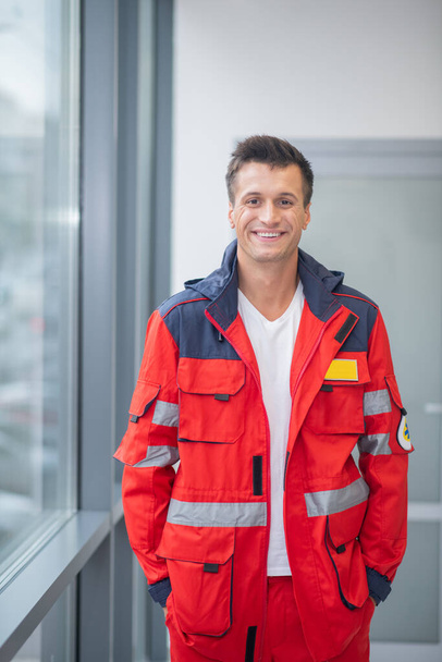 Male doctor in red uniform standing near the window and smiling - Fotoğraf, Görsel