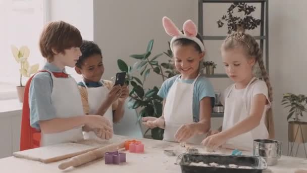 Waist up of Caucasian and Mixed-Race children playing with flour and dough, standing at kitchen table, African boy taking pictures of them with smartphone. Diverse kids in aprons having time together - Footage, Video