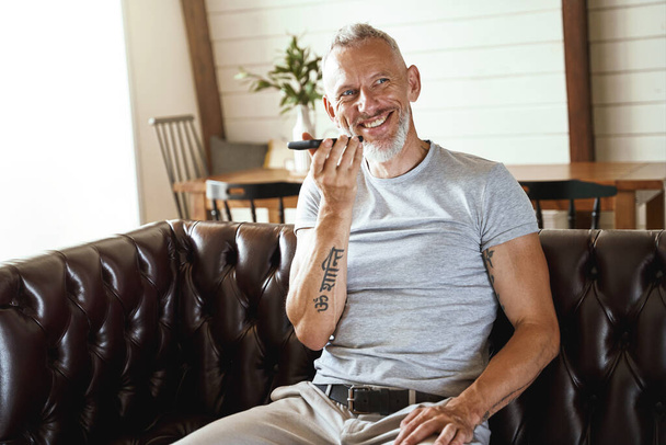 Onnellinen keski-ikäinen mies rento vaatteet puhuu matkapuhelimella ja hymyilee istuessaan sohvalla olohuoneessa, työskentelee etänä - Valokuva, kuva