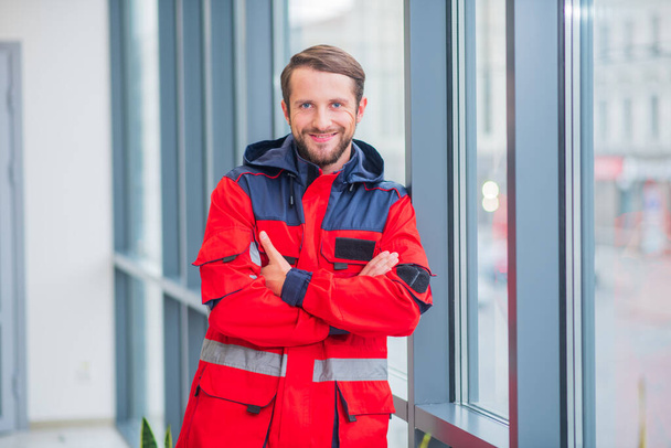 Bearded doctor in red uniform standing near the window - Фото, изображение
