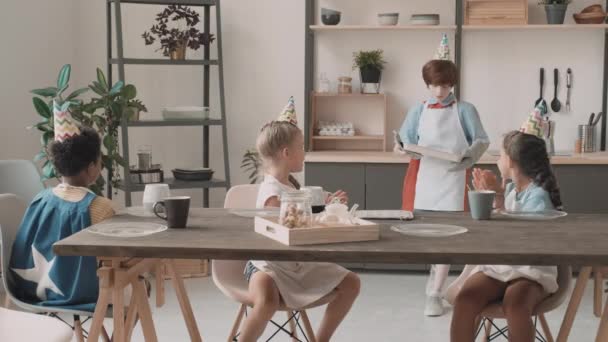 Średni długi rudowłosy biały chłopiec serwujący gorące ciasteczka na blasze do różnych przyjaciół siedzących przy kuchennym stole, klaszczących w dłonie. Wielokulturowe dzieci w kapeluszach i fartuchach imprezowych - Materiał filmowy, wideo