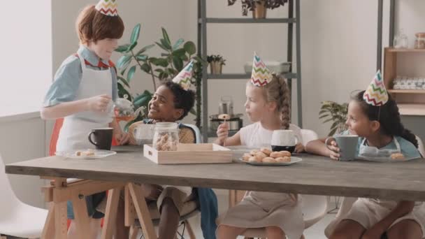 Menino africano, meninas caucasianas e mestiças sentadas à mesa de jantar, comendo biscoitos, cara da escola de cabelos vermelhos derramando chá para os amigos. Crianças diversas usando aventais e chapéus de festa, sorrindo - Filmagem, Vídeo