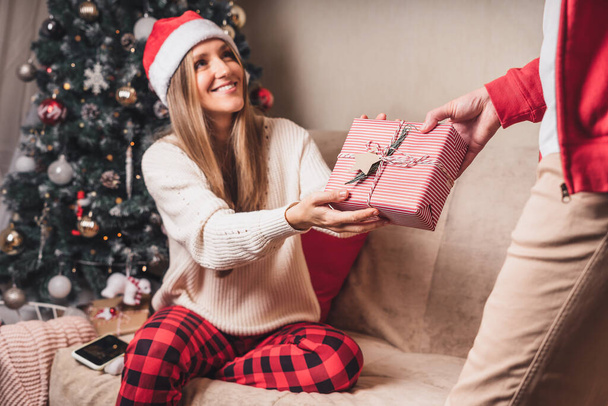 Vista trasera de cerca del hombre en suéter rojo sosteniendo una caja de regalo de Navidad a sus espaldas mientras está de pie cerca del árbol de Navidad decorado. - Foto, Imagen