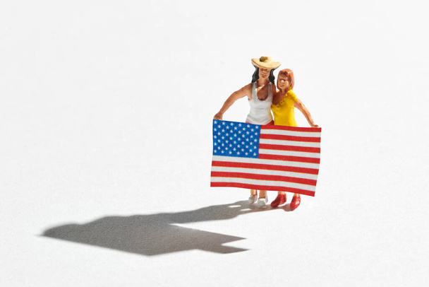 Miniatur zweier amerikanischer Frauen mit Fahne. Demonstrationsteilnehmer, politische Wahlkampfunterstützer. Hochwinkelansicht isoliert auf weißem Hintergrund mit Schatten und Kopierraum - Foto, Bild