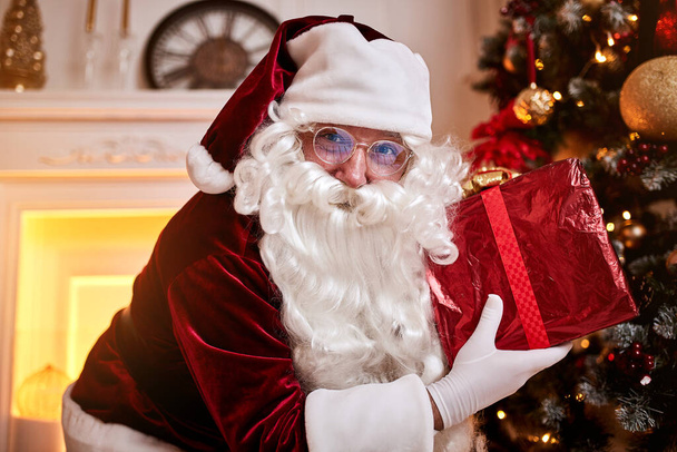 Szczęśliwy Święty Mikołaj przyniósł pudełko prezentów dla dzieci w pobliżu pięknej choinki i kominka. Nowy rok i Wesołych Świąt koncepcja - Zdjęcie, obraz