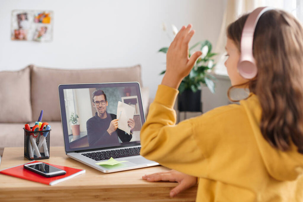 School girl is studying remotely, using laptop webcam and headphones, raises her hand to answer teacher's question. Home schooling and distance education concept. Online tutor having distance lesson - Photo, Image