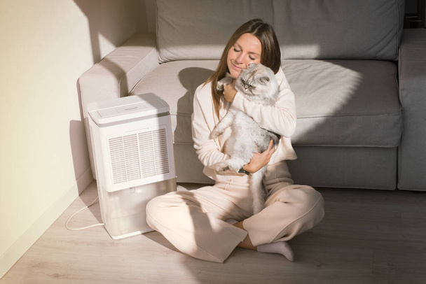 Mulher com o gato a respirar ar fresco em casa. Purificador de ar ou umidificador em uma sala de estar. Desumidificador, indicador de umidade, ionizador de ar ou recipiente de água. - Foto, Imagem