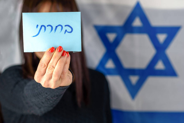Young Woman Holding Ballot Front Of Face on Israeli Flag Background.Hebrew text I Voted on voting paper. - Foto, Bild