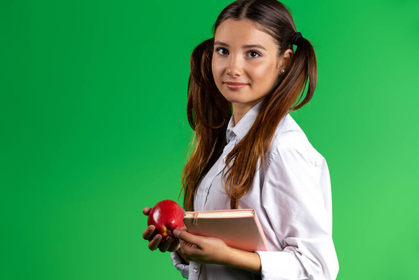 A schoolgirl in a school white shirt, holding an apple in one hand and a book in the other. Posing against a green background. High quality photo - Foto, Bild