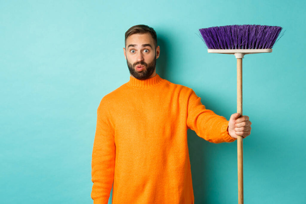 Shocked man receiving broom to do house chores, looking confused, standing over light blue background - Foto, immagini