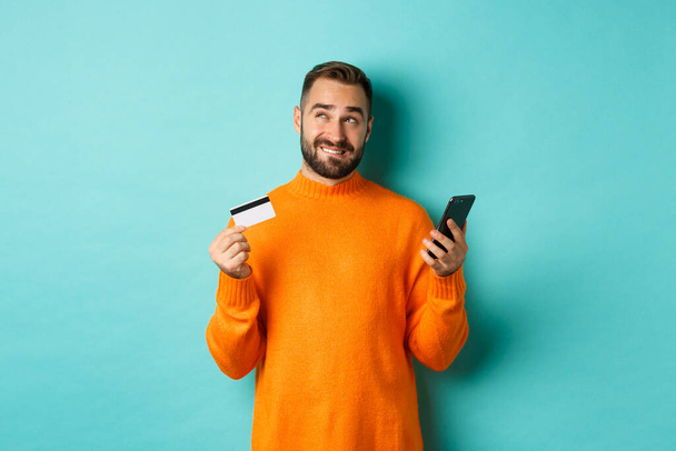 Compras en línea. Hombre reflexivo que sostiene la tarjeta de crédito y el teléfono móvil, pensando en la compra, de pie sobre fondo azul claro - Foto, imagen