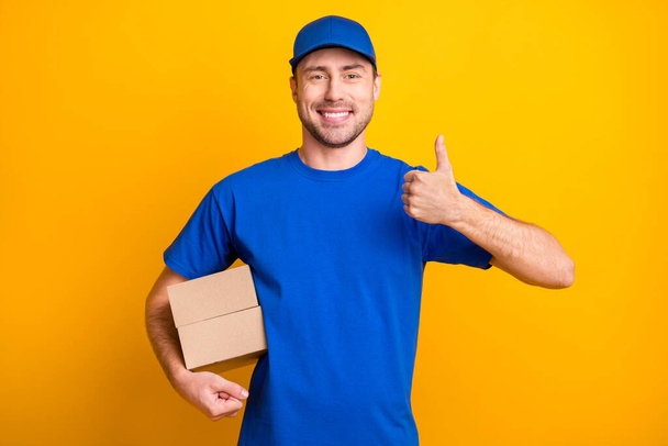 Photo of positive guy show thumb up arm hold carton package wear hat isolated on yellow color background - Foto, Bild