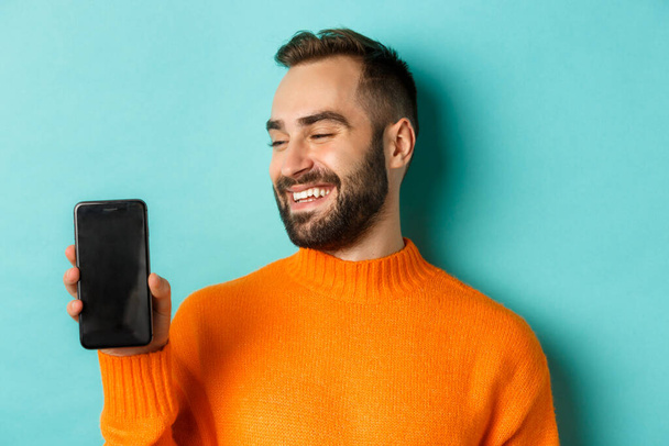 Primer plano del joven barbudo mostrando la pantalla del teléfono y mirando satisfecho, usando suéter naranja, de pie sobre el fondo del estudio - Foto, imagen
