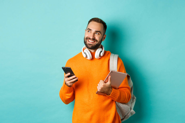 Handsome man student with headphones and backpack, holding digital tablet and smartphone, looking dreamy at upper left corner, standing against turquoise background - Fotoğraf, Görsel