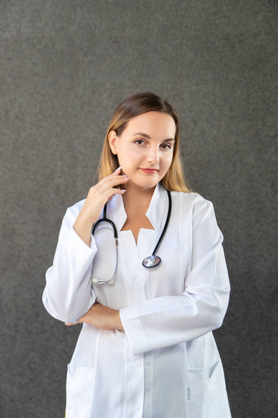 Portrait of a modern doctor's girl standing isolated on a gray background with a stethoscope, holding her hand to her face. Studio, copy space. Medicine, healthcare and profession concept. - Fotografie, Obrázek