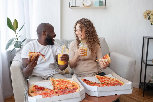 Delicious lunch break on the sofa in the living room. Happy young couple look joyfully at each other, eating pizza. Family, pregnancy, happiness and food concept. - Photo, Image