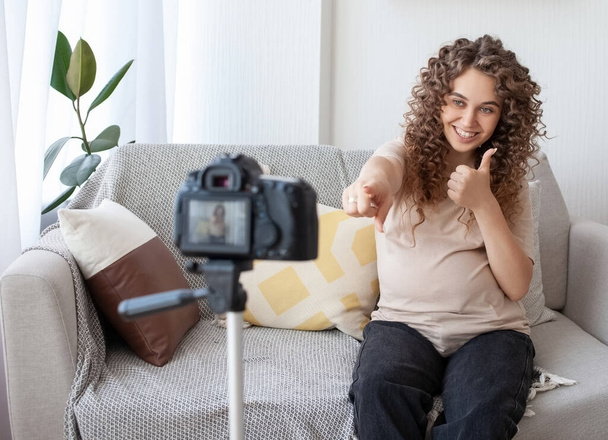 Cute curly future mom shoots video for her blog with camera and tripod, gesticulates and invites everyone to subscribe to her channel. People, pregnancy and blogging concept. - Fotó, kép