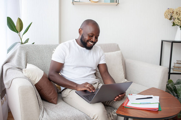 Hymyilevä afrikkalainen mies makuuhuoneessa istuu harmaalla sohvalla tekee bisnestä, tehden videoneuvottelua kollegoidensa kanssa. Pääkonttori. Liiketoiminnan, viestinnän, ihmisten ja teknologian käsite. - Valokuva, kuva