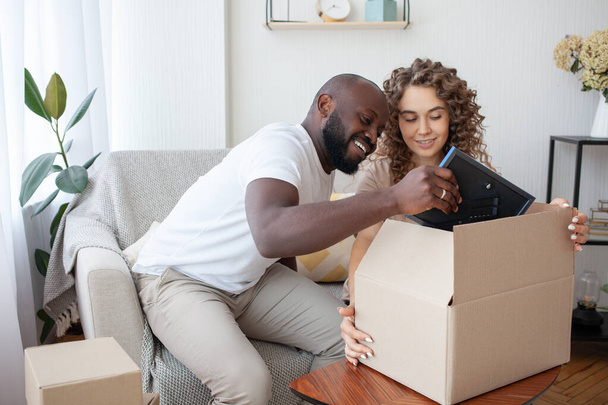 Happy married multinational couple packing a package for their relatives, put their picture in a cardboard box in a blue frame. Family, relationships and happiness concept. - Photo, Image