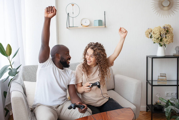 Active rest of a young wonderful couple. African-American and expectant mother celebrate draw in video game, raising their hands up and holding joysticks. Technology, family and pregnancy concept. - Photo, Image