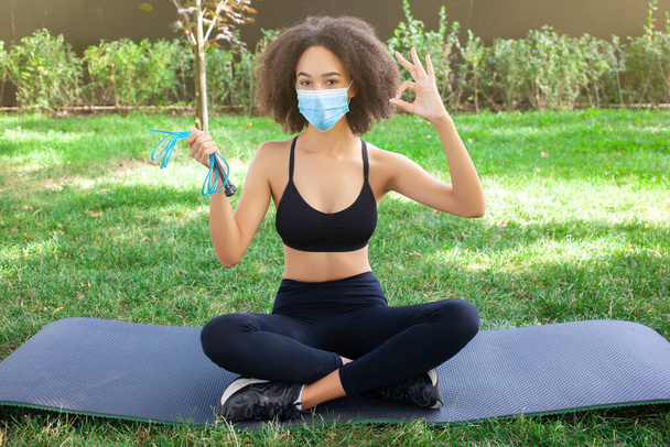 A sportos afro-amerikai lány maszkot visel, jól érzi magát és nyugodt, kötelet tart a közelgő edzéshez a kertben, divatos fekete sportruhába öltözve. Egészségügyi, karantén- és sportkoncepció. - Fotó, kép