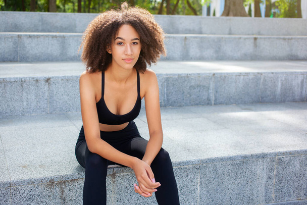 若いアフリカ系アメリカ人女性のランナーの写真は、公園でのスポーツ活動の後に休憩し、階段の上の黒いトレンディーなトラックスーツで走ります。スポーツ、アウトドア、健康的なコンセプト. - 写真・画像