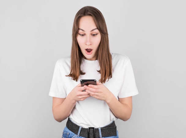 Portrait if a shocked young girl looking at mobile phone isolated over gray background - Φωτογραφία, εικόνα