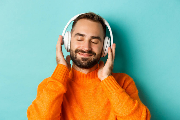 Close-up of handsome modern man listening music in headphones, standing in orange sweater over turquoise background - 写真・画像
