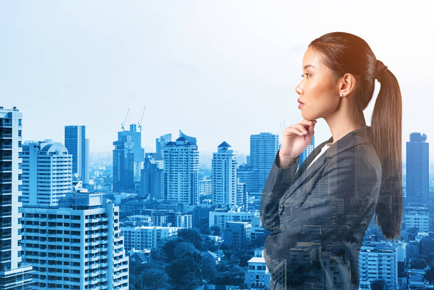 Attractive young Asian business woman in suit with hand on chin thinking how to succeed, new career opportunities, MBA. Bangkok on background. Double exposure. - Photo, Image