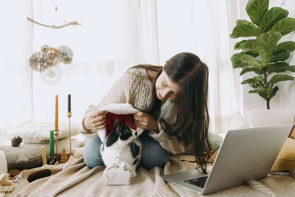 Stylish woman in cozy sweater putting santa hat on cute cat at christmas furoshiki gift and laptop in festive decorated boho room. Young female playing with furry friend. Happy Holidays! - Фото, зображення