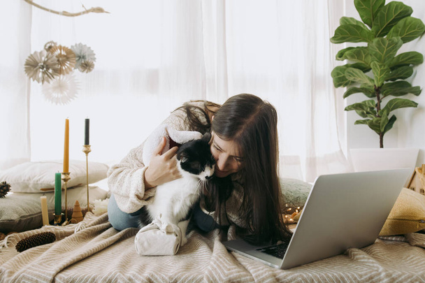 Stylish woman in cozy sweater caressing and kissing cute cat in  santa hat at christmas furoshiki gift and laptop in festive decorated boho room. Celebrating at home, Merry Christmas! - Photo, Image