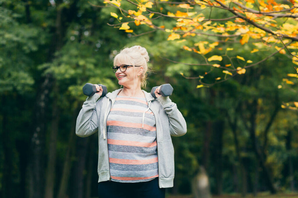 Hermosa mujer mayor en un paseo por el parque, realiza ejercicios con mancuernas en sus manos - Foto, imagen