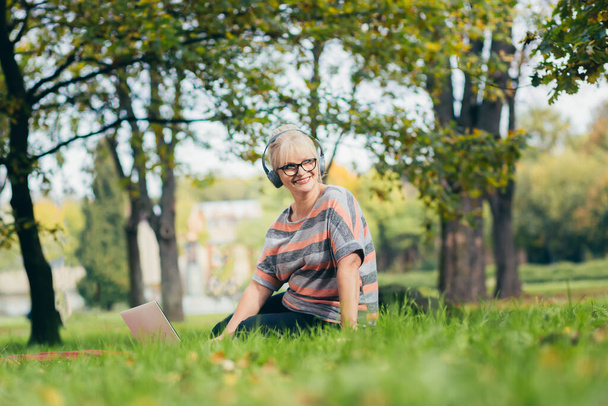 Senior woman resting in the park sitting on the grass, using phone and laptop, and reading a book - 写真・画像