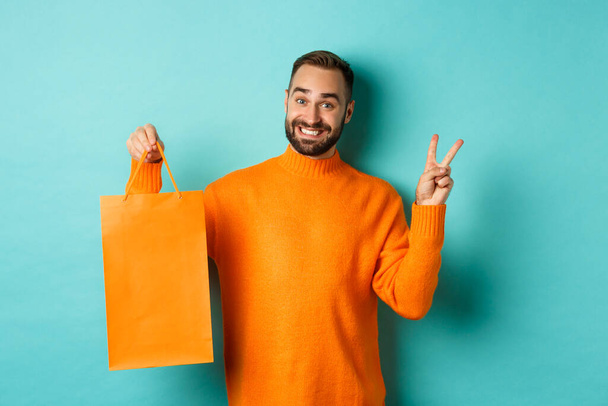 幸せな若者が平和サインとオレンジのショッピングバッグを見せ、満面の笑みを浮かべて、ターコイズの背景に立って - 写真・画像