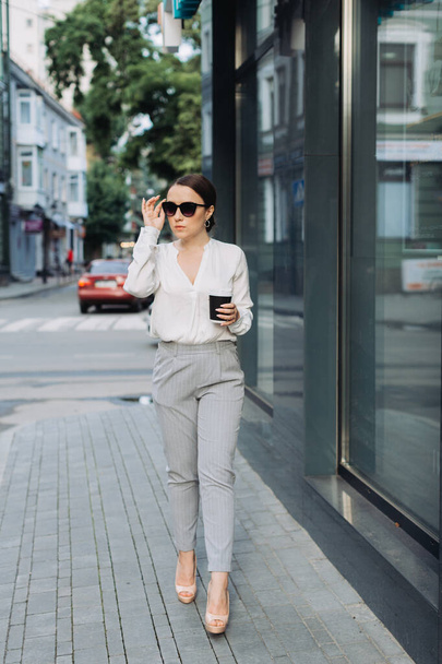 路上でコーヒーを飲みながら携帯電話で笑顔の若いビジネス女性のショット. - 写真・画像