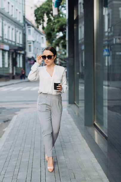Shot van glimlachende jonge zakenvrouw met behulp van haar mobiele telefoon tijdens het drinken van koffie lopen door de straat. - Foto, afbeelding