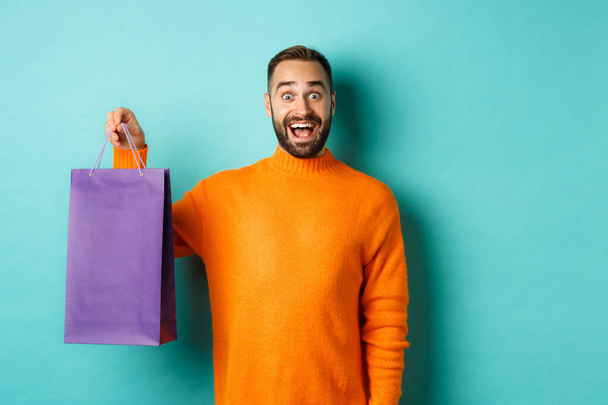 Excited adult man holding purple shopping bag and smiling, going to mall, standing over turquoise background - Foto, imagen